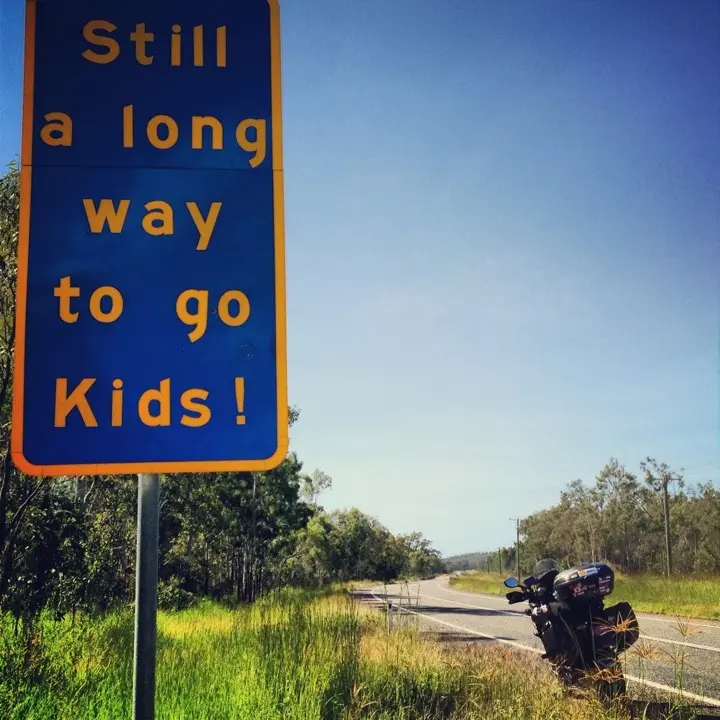 A roadside sign reading Still a long way to go kids beside an open Queensland road